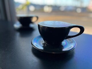 Low angle view two coffee cups, focus on foreground cup, with background street, through window, in soft focus
