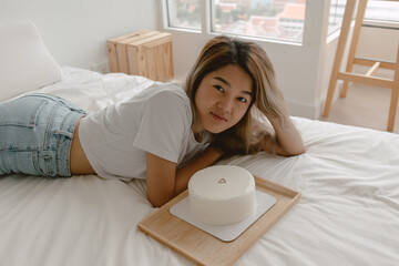 Lovely beautiful happy asian woman lies on the bed admiring her birthday cake.