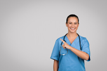 Positive caucasian female doctor surgeon in blue uniform pointing finger at copy space on gray...