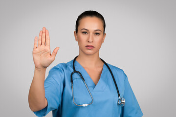 Serious european female doctor in blue uniform showing stop gesture, refusing something, saying no...