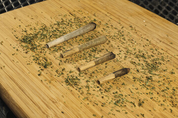Marijuana cigarettes of different sizes on a wooden board, with remains of ground marijuana around.