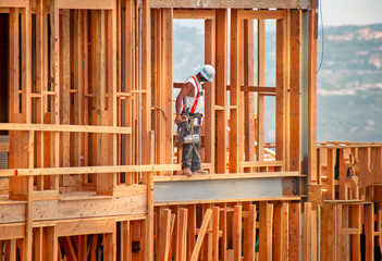 Horizontal image of a construction worker standing on wood framing wearing a tool belt, hard hat and safety harness. The shot is in afternoon light