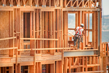 Horizontal image of a construction worker standing on wood framing wearing a tool belt, hard hat and safety harness. The shot is in afternoon light and the worker is in the right side of the frame.