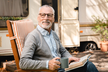 Closeup photo of old senior elderly grandfather man adventurer explorer relaxing in a chair reading book while traveling caravanning in motor home camper van trailer - obrazy, fototapety, plakaty