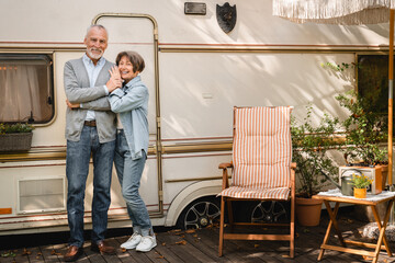 Full length photo of confident caucasian senior old travelers adventurers couple spouses hugging embracing looking at camera while traveling by trailer camper van motor home