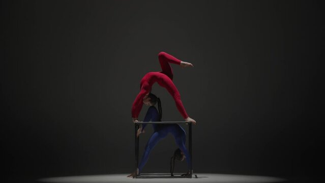 Portrait of two female acrobats isolated on black background. Girls aerial dancers in blue red suits performing bridge on a cube.