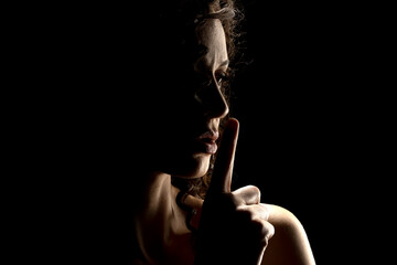 Silhouette of unknown woman with the face in the shadow holding finger on her lips on a black...