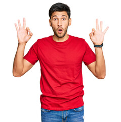 Fototapeta premium Young handsome man wearing casual red tshirt looking surprised and shocked doing ok approval symbol with fingers. crazy expression