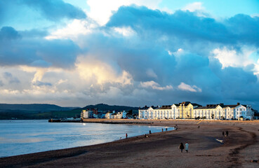 A beach in golden panoramic landscape view. Orange dusk sunlight is cast across the beach and ornate white buildings. People walking on the beach.