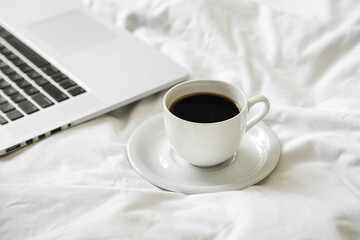 Cup of black coffee, laptop and smartphone on unmade white bed, work from home concept.