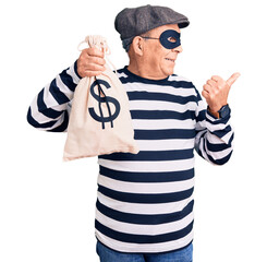 Senior handsome man wearing burglar mask holding money bag pointing thumb up to the side smiling...