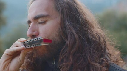 the musician enjoys playing the harmonica while standing on the street. a young man with long hair plays the harmonica.