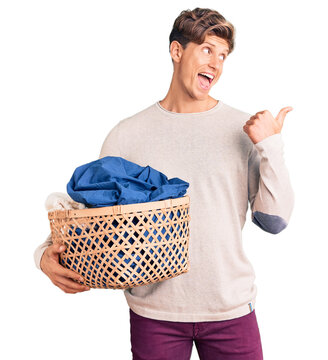 Young handsome man holding laundry basket pointing thumb up to the side smiling happy with open mouth