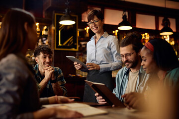 Happy waitress using touchpad while talking to guests in bar.