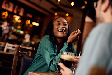 Cheerful black woman has fun while talking to her friend and drinking beer in bar.