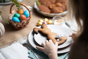 Hands of child decorating bunny shaped gingerbread cookies with icing on festive decorated table on Easter. Holiday traditions, family time - obrazy, fototapety, plakaty