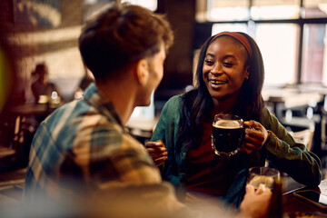 Happy black woman communicating with her friend while enjoying in glass of beer in pub. - obrazy, fototapety, plakaty