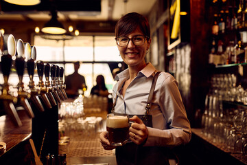 Happy waitress pouring beer draft beer at bar counter and looking at camera.