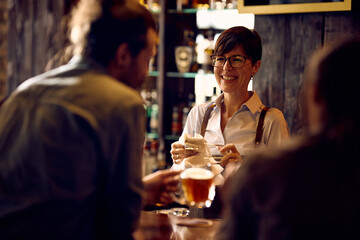 Happy waitress communicating with customers while cleaning glasses at bar counter.