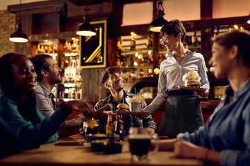 Happy waitress serving food to group of friends in pub.