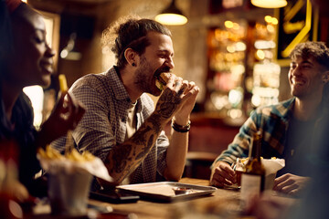 Young man eating burger while being with friends in pub. - obrazy, fototapety, plakaty