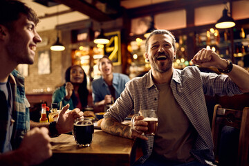 Young man and his friends cheering while watching sports game in bar.