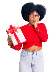 Young african american girl holding gift afraid and shocked, surprise and amazed expression with hands on face