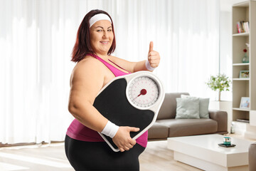 Plus size woman in sportswear holding a weight scale in a living room at home