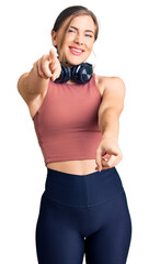 Beautiful caucasian young woman wearing gym clothes and using headphones pointing to you and the camera with fingers, smiling positive and cheerful