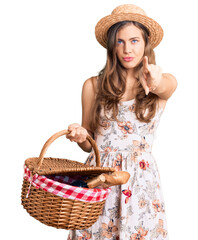 Beautiful caucasian young woman wearing summer hat and holding picnic wicker basket with bread pointing with finger to the camera and to you, confident gesture looking serious