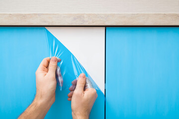 Young adult man hands removing blue protective polythene film from new kitchen cabinet doors under table top. Closeup. Front view.