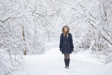 Young adult woman walking on fresh white snow covered road through tree branches at park after blizzard in beautiful cold winter day. Spending time alone. Front view. Peaceful atmosphere in nature.