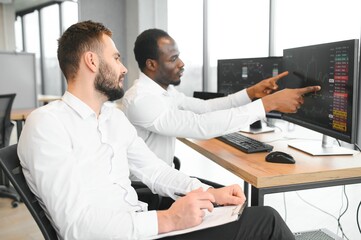 Two successful trader sitting in office, checking cryptocurrency information data on finance market graph, pointing on monitor