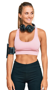 Young blonde woman wearing gym clothes and using headphones looking away to side with smile on face, natural expression. laughing confident.