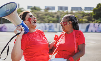 Mature latin female friends football supporter fans watching soccer match event at stadium - Senior women with red t-shirts having excited fun on sport world championship