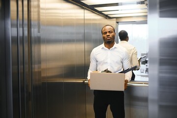 A young african man fired from work is standing in the elevator with a box of things