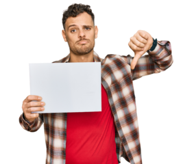 Young hispanic man holding blank empty banner with angry face, negative sign showing dislike with thumbs down, rejection concept