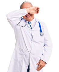 Senior handsome grey-haired man wearing doctor coat and stethoscope covering eyes with arm smiling cheerful and funny. blind concept.