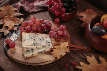 Cheese platter served with autumn fruits and dry leaves on dark wooden background. Appetizer of blue cheese on cutting board with grapes