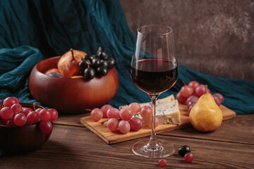 Glass of rich red wine served with cheese plate and fruits on dark wooden background. Appetizers table with wine glasses, rustic style table setting in winery