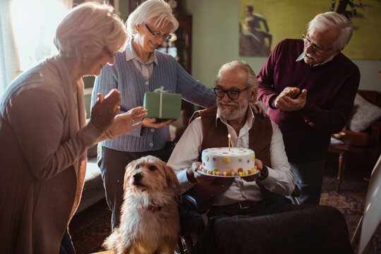 Senior friends celebrating birthday with cake and gift at home with a dog
