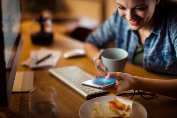 Woman smiling and using smartphone while having breakfast at home computer