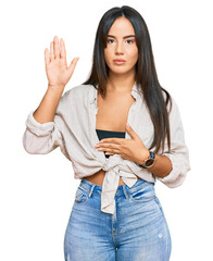 Young beautiful hispanic girl wearing casual clothes swearing with hand on chest and open palm,...