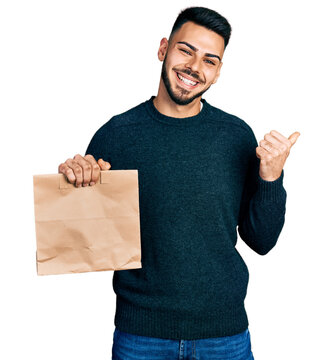 Young hispanic man with beard holding take away paper bag pointing thumb up to the side smiling happy with open mouth