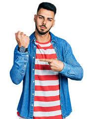 Young hispanic man with beard wearing casual denim jacket in hurry pointing to watch time,...