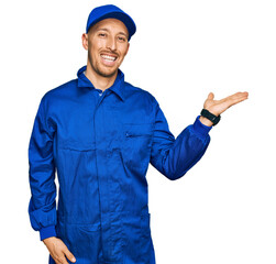 Bald man with beard wearing builder jumpsuit uniform smiling cheerful presenting and pointing with palm of hand looking at the camera.