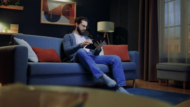Man working at home. Male sitting at blue couch. Man holding tablet and touching screen. Male wearing glasses and working online. Man relaxing at home. Man looking at tablet.