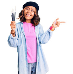 Young beautiful mixed race woman holding paintbrushes smiling happy pointing with hand and finger...