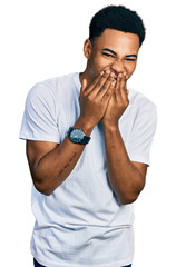 Young african american man wearing casual white t shirt laughing and embarrassed giggle covering mouth with hands, gossip and scandal concept
