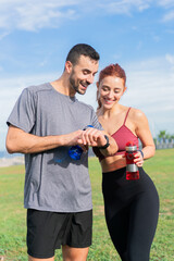 Two sporty young adults resting and checking the smart band analyzing the workout smiling and laughing together outdoors. Two people fitness resting with water bottles and with sportswear